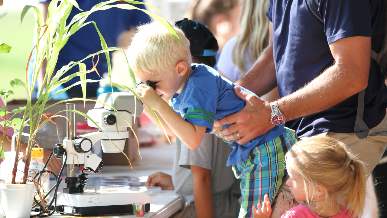 Young boy looking into a microscope, held up by father with daughter at Auburn's Ag Roundup event.