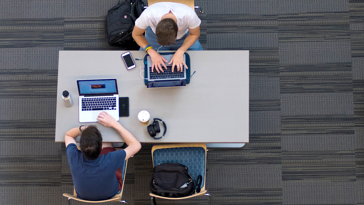 Two Auburn university students study on laptops at library desk with backpacks, student resources