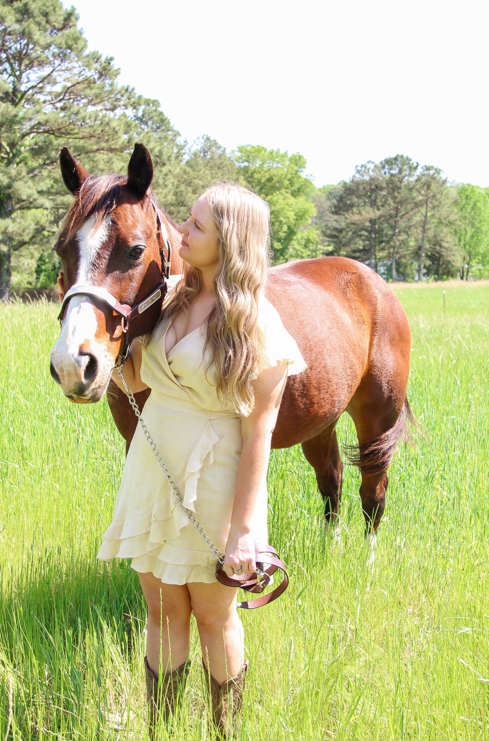 A blonde young woman in a cream colored dress holds the reins of a brown horse with white markings on its face in a field of green grass
