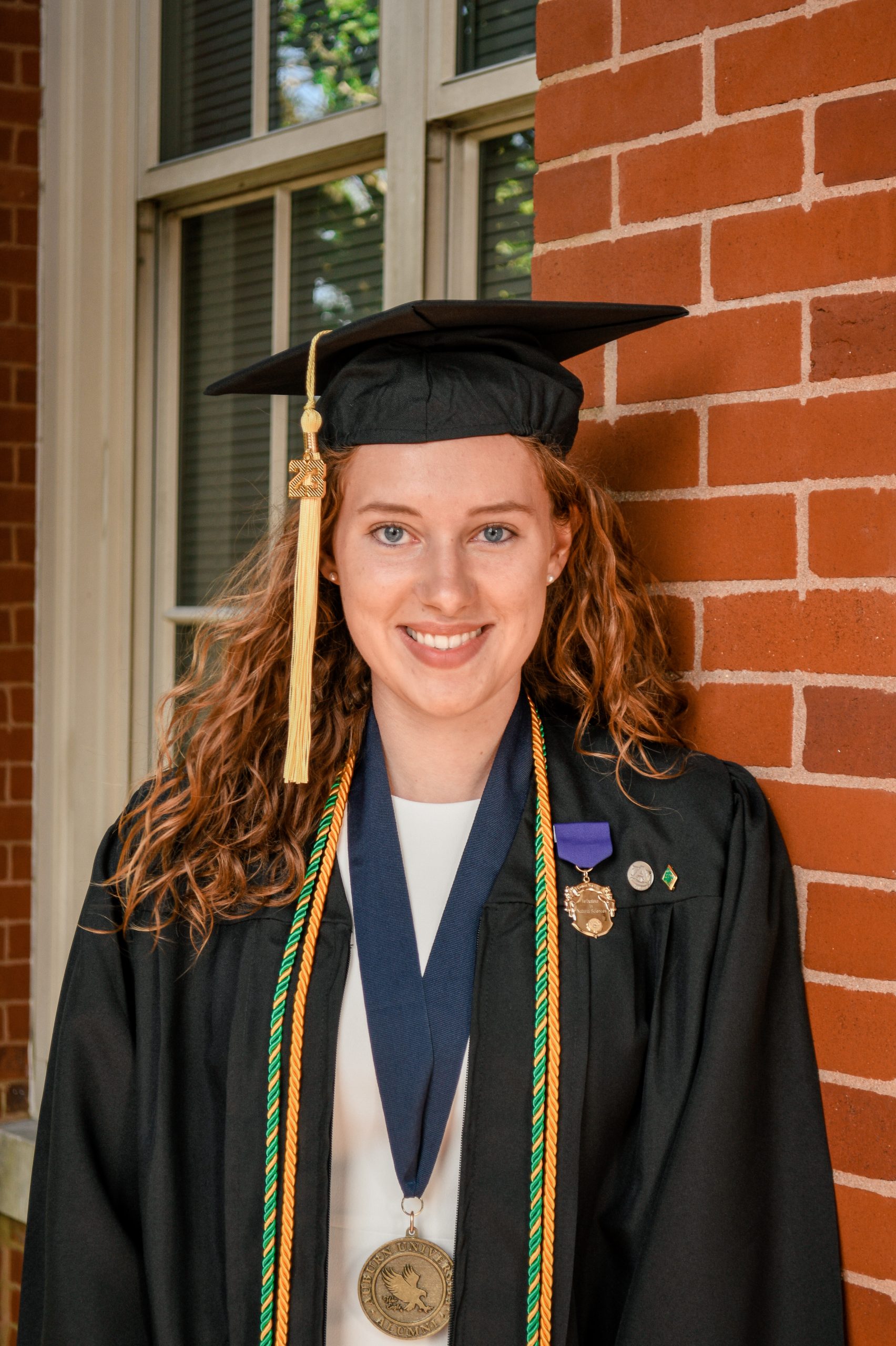 A white young woman with curly red hair wears a cap and gown and a medal around her neck while she leans against a red-brick wall with a window behind her