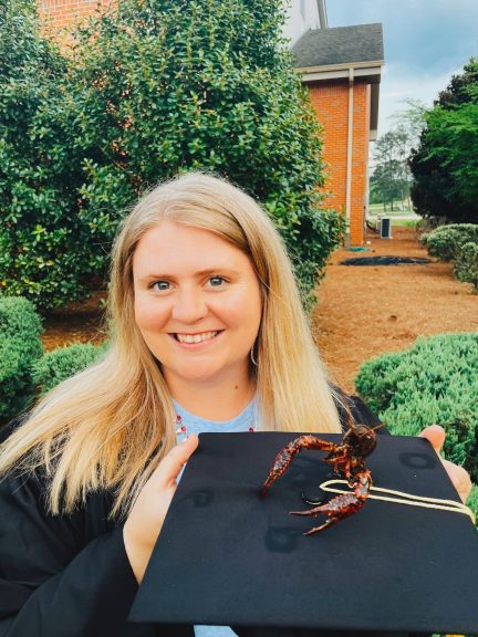 A blond woman holds a graduation cap with a crawfish on it in front of a tall green bush with a red-orange brick building behind it