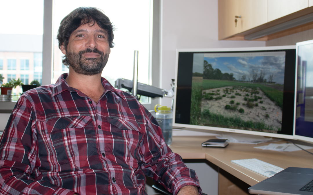 A bearded, brown-haired man in a red plaid shirt smiles while seated at a desk in front of a computer with an image of cover crops on it.