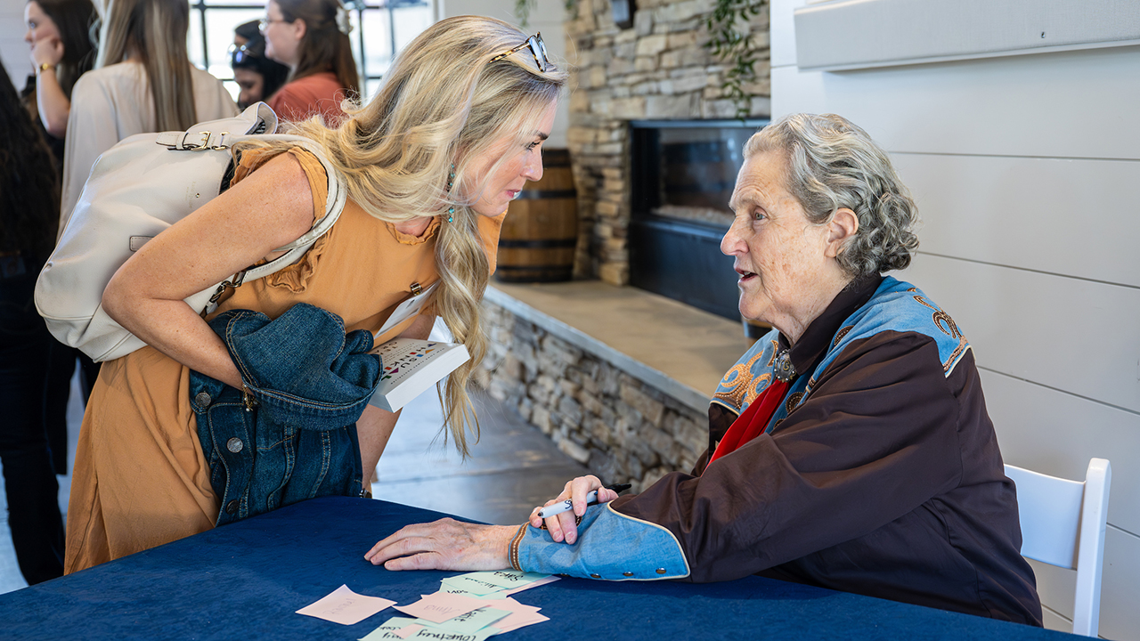 a blond woman in an orange dress with a handbag over her shoulder leans over a table to speak to Temple Grandin, who is seated