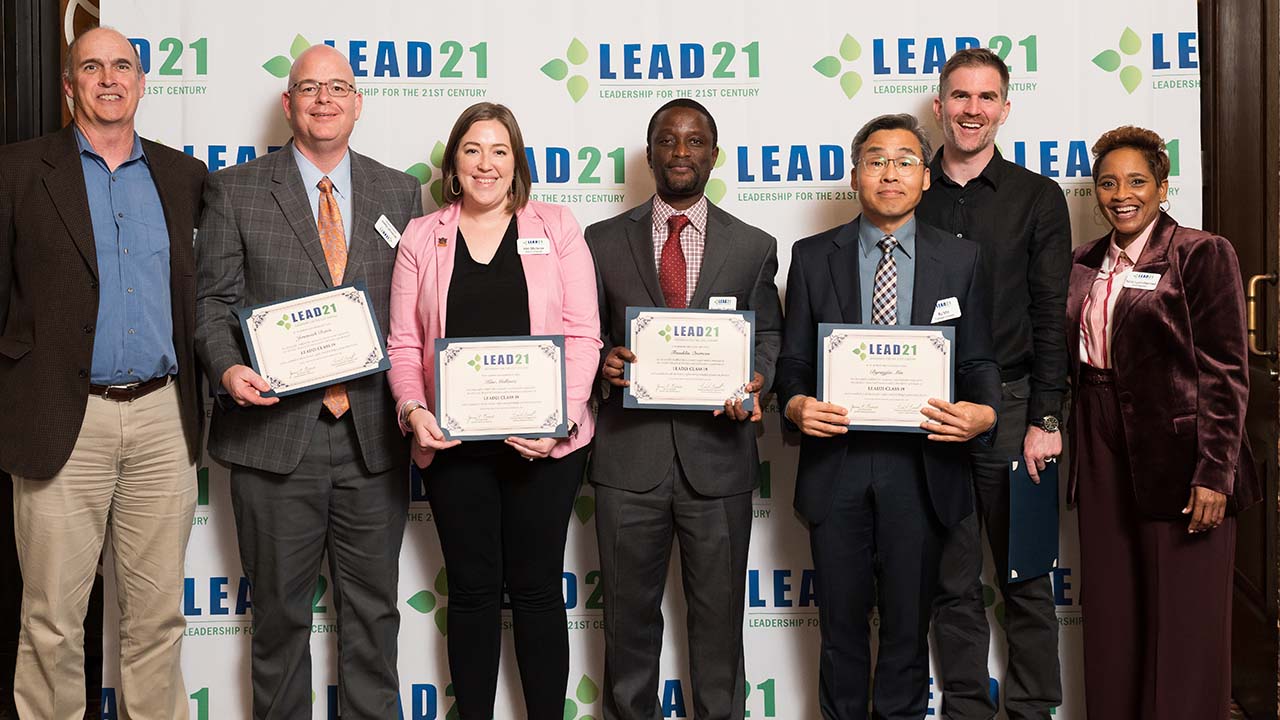 Seven individuals, men and women, pose with certificates in front of a LEAD21 backdrop.